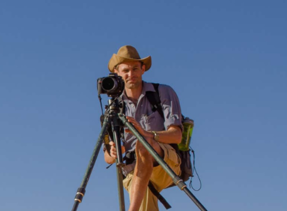 A man wearing a hat and shorts sits comfortably on a tripod