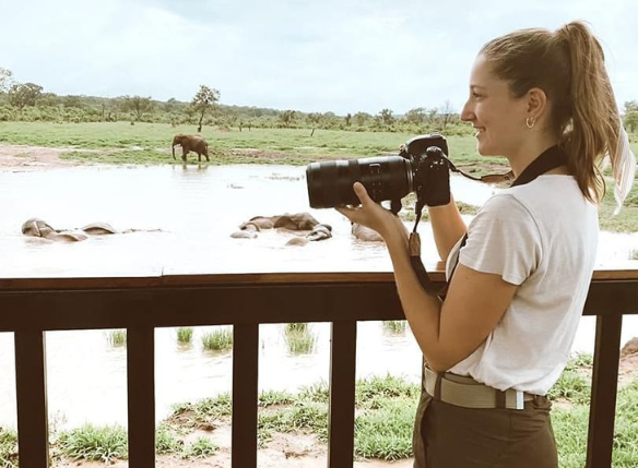 hanna wigart photographing holding her camera with a water hole and elephant in the background