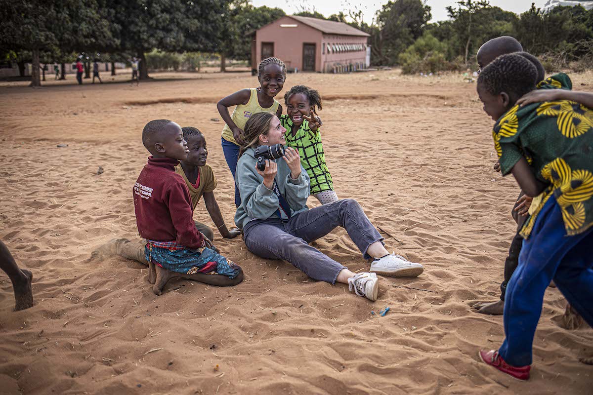 A woman with a camera sits on the ground, surrounded by smiling children