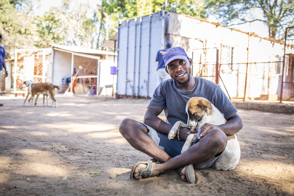 A man sitting on the ground smiles while holding a dog in his lap