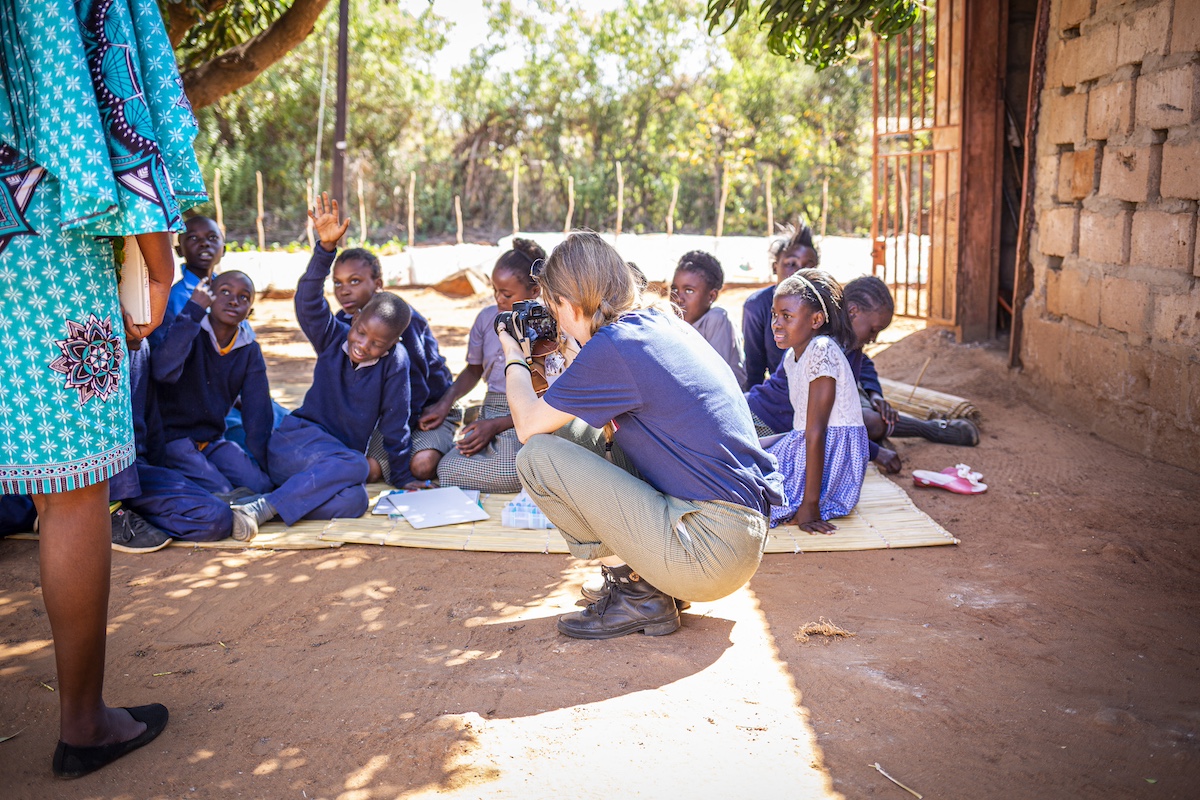 A woman crouches with a camera, photographing a group of children sitting on the ground