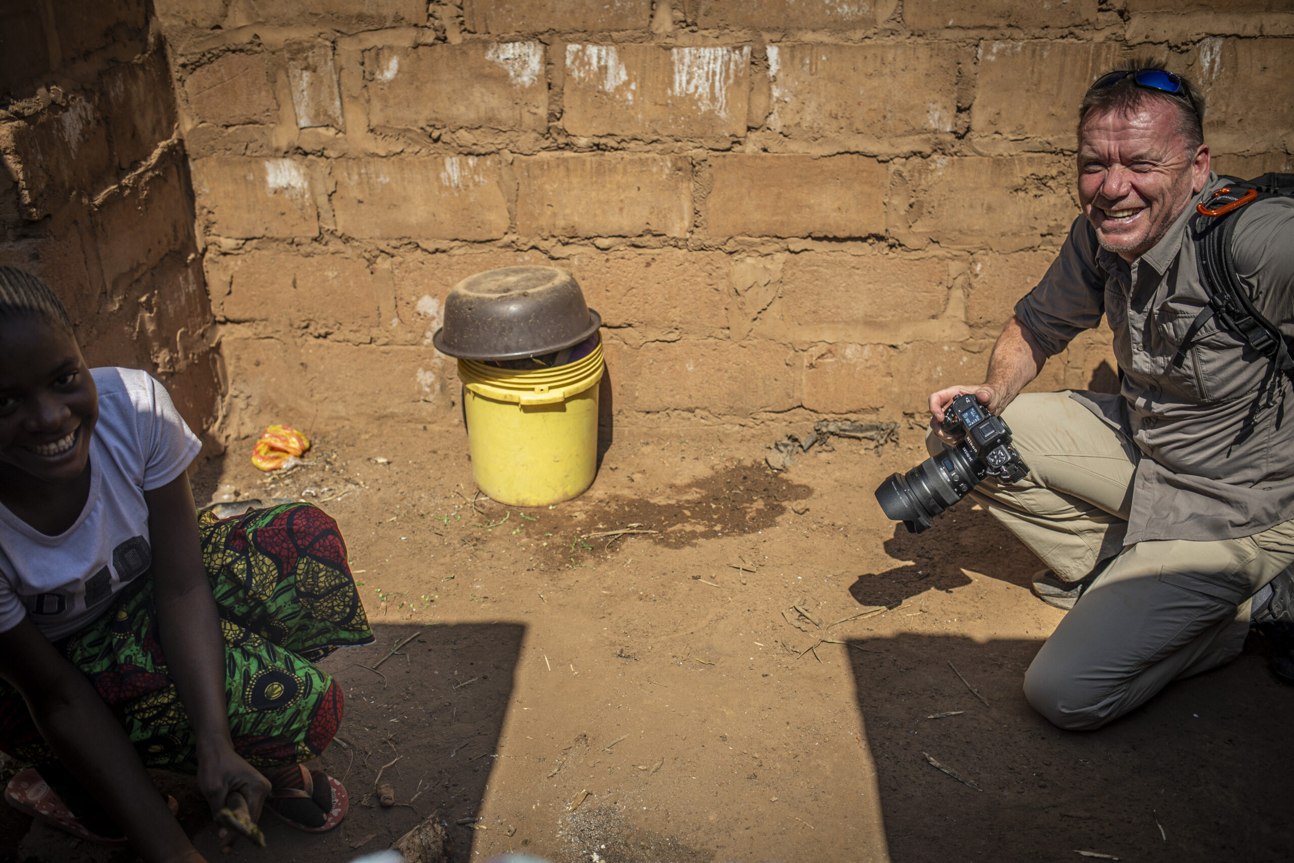 A man with a camera crouches and smiles beside a woman seated