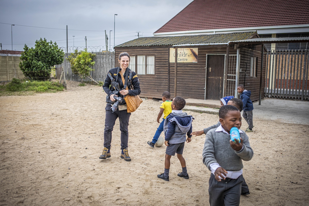 A woman holding a camera stands outside near a group of children playing