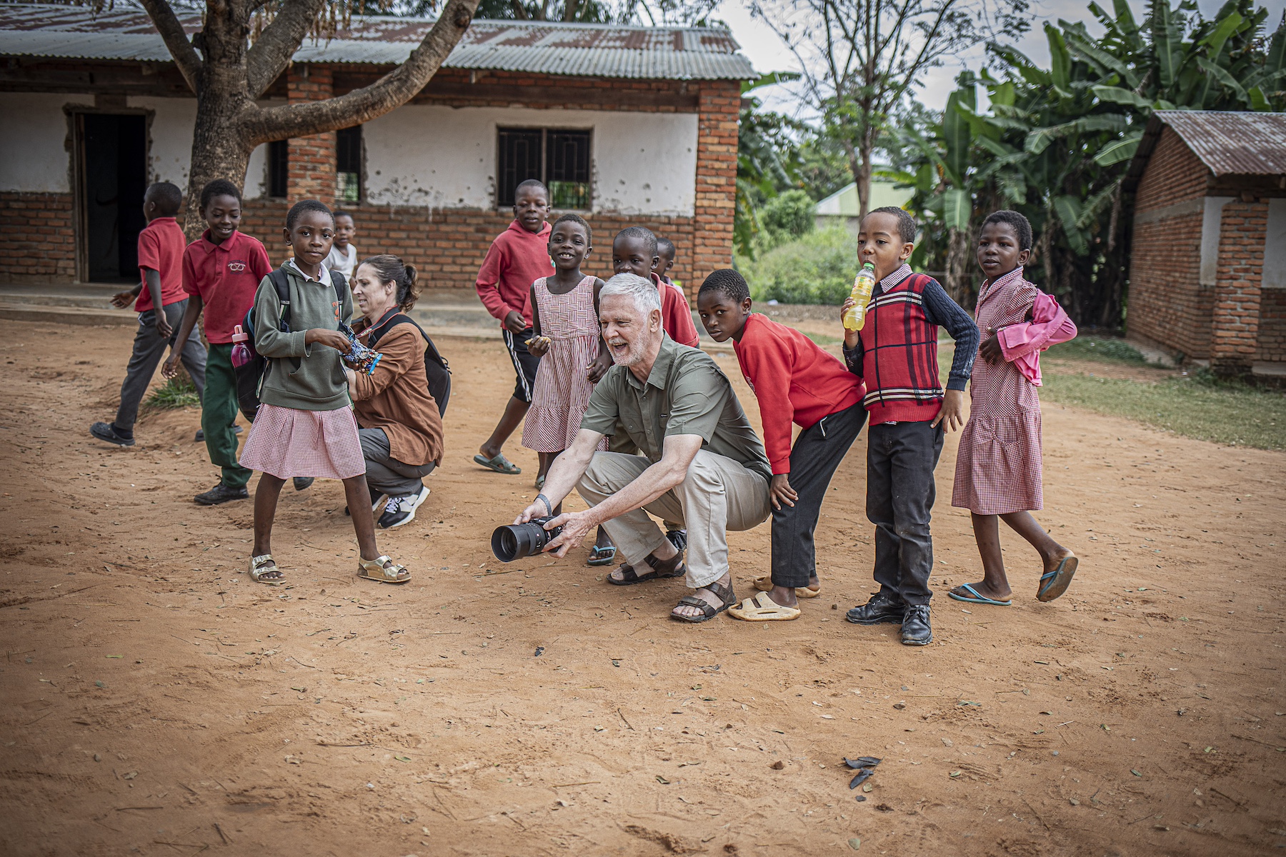 A group of children and two adults outdoors, with one adult crouching and holding a camera, surrounded by children in a village