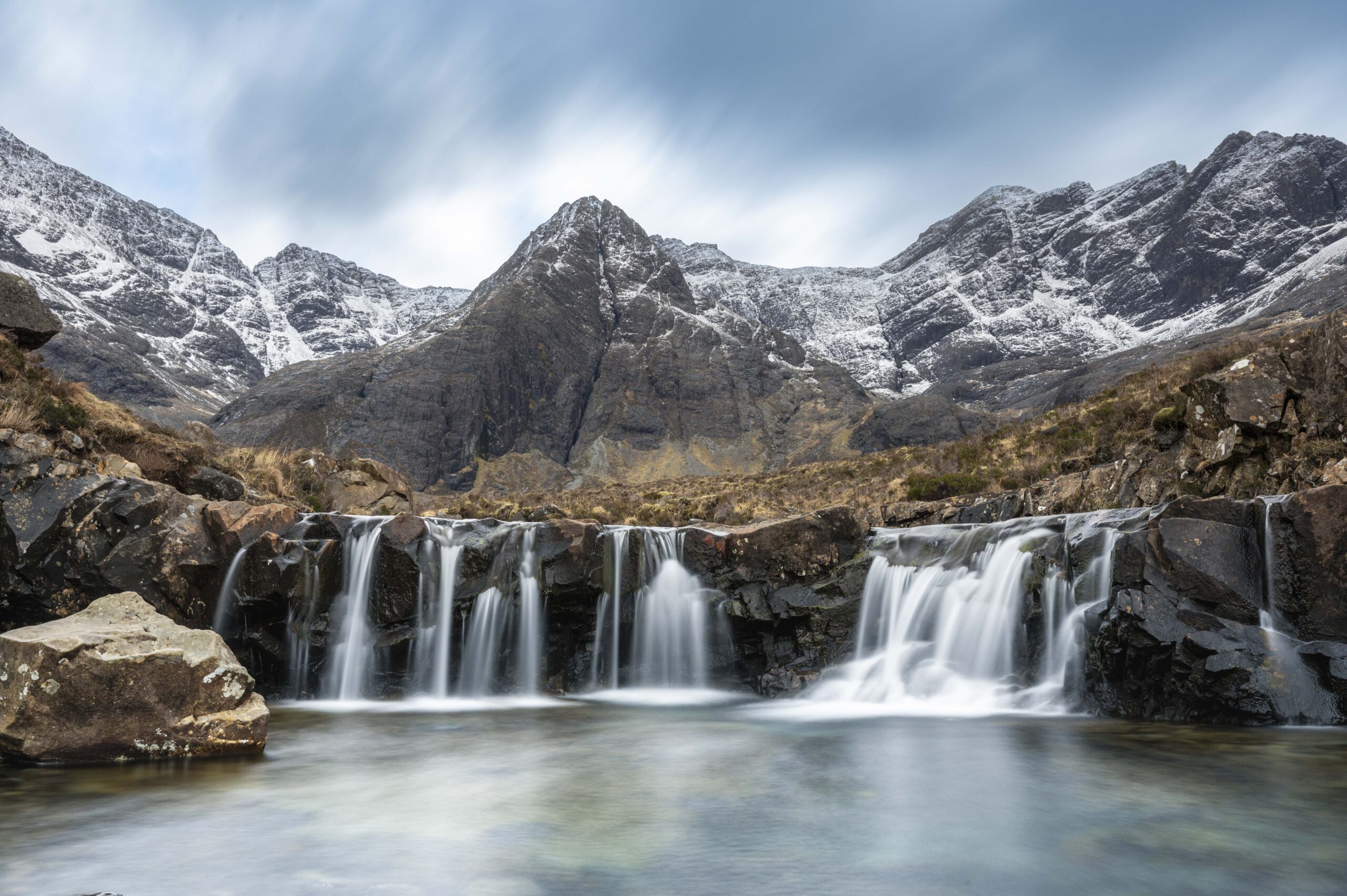 Small waterfalls flowing into clear pools with snow-covered mountains in the background