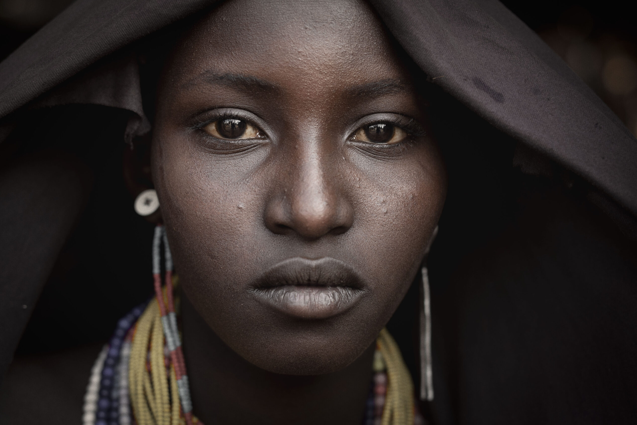 A close-up portrait of a woman with a dark hood and colorful beaded necklace