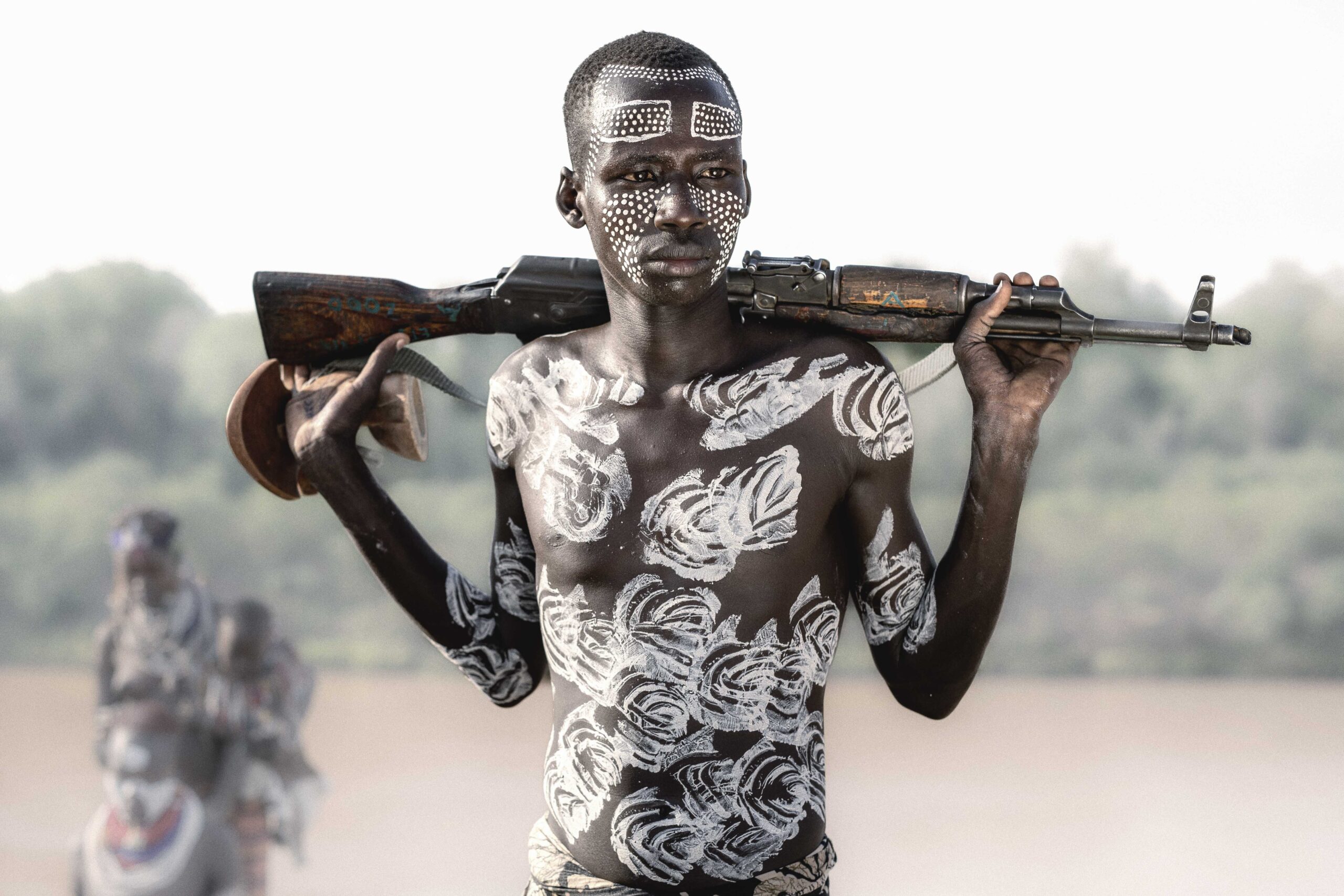 A young man with body paint and face markings holding a rifle across his shoulders