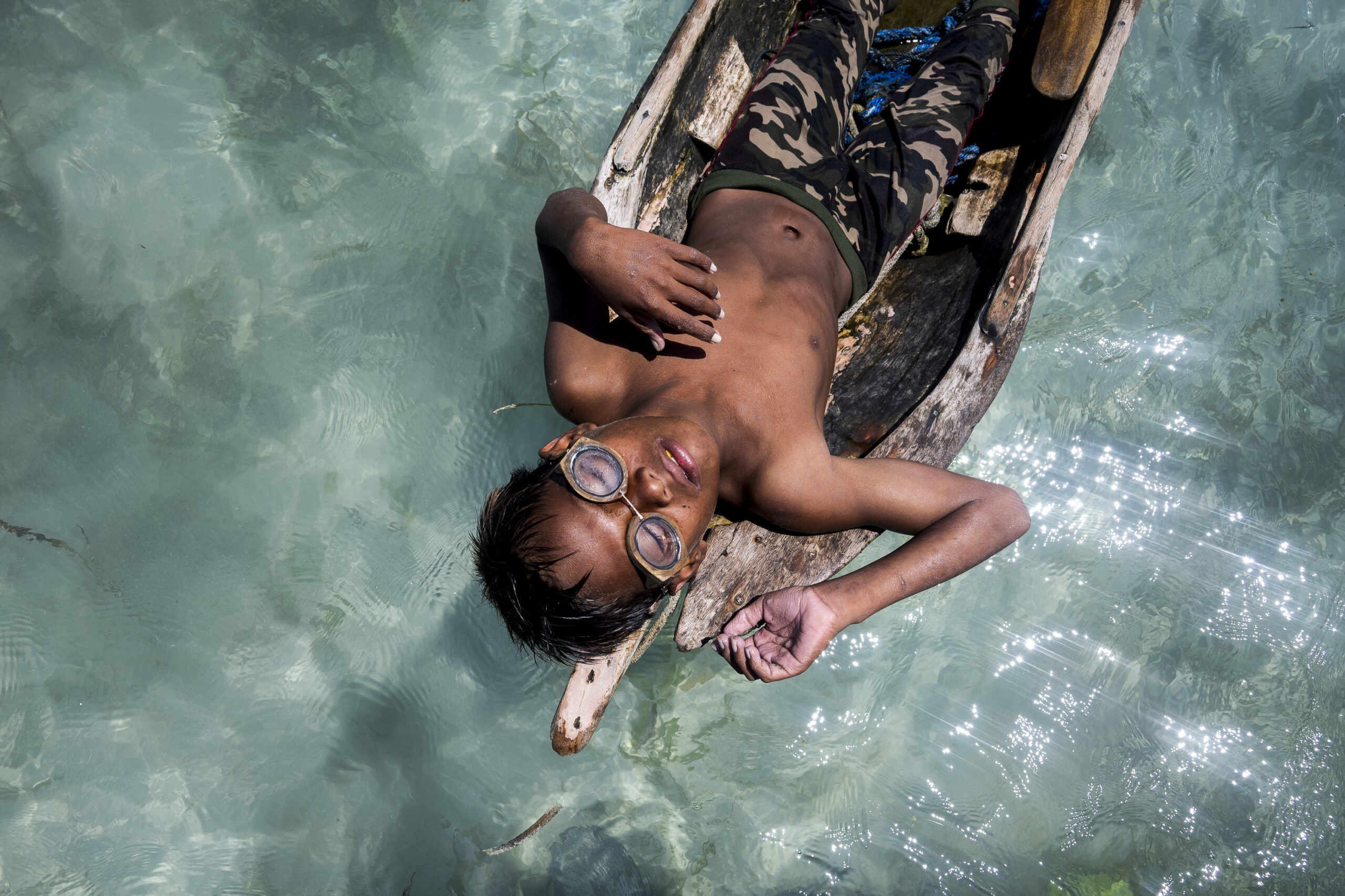 A boy wearing goggles lying in a wooden boat on clear turquoise water