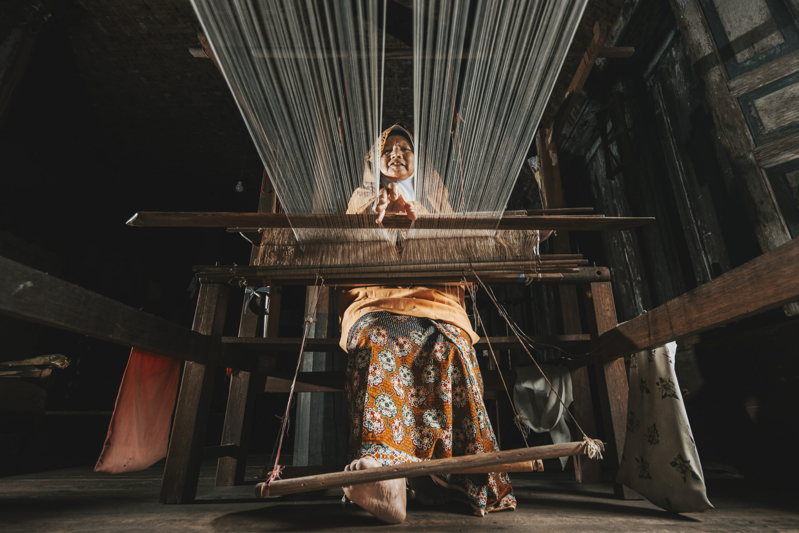 A woman weaving fabric on a traditional loom in a dimly lit room