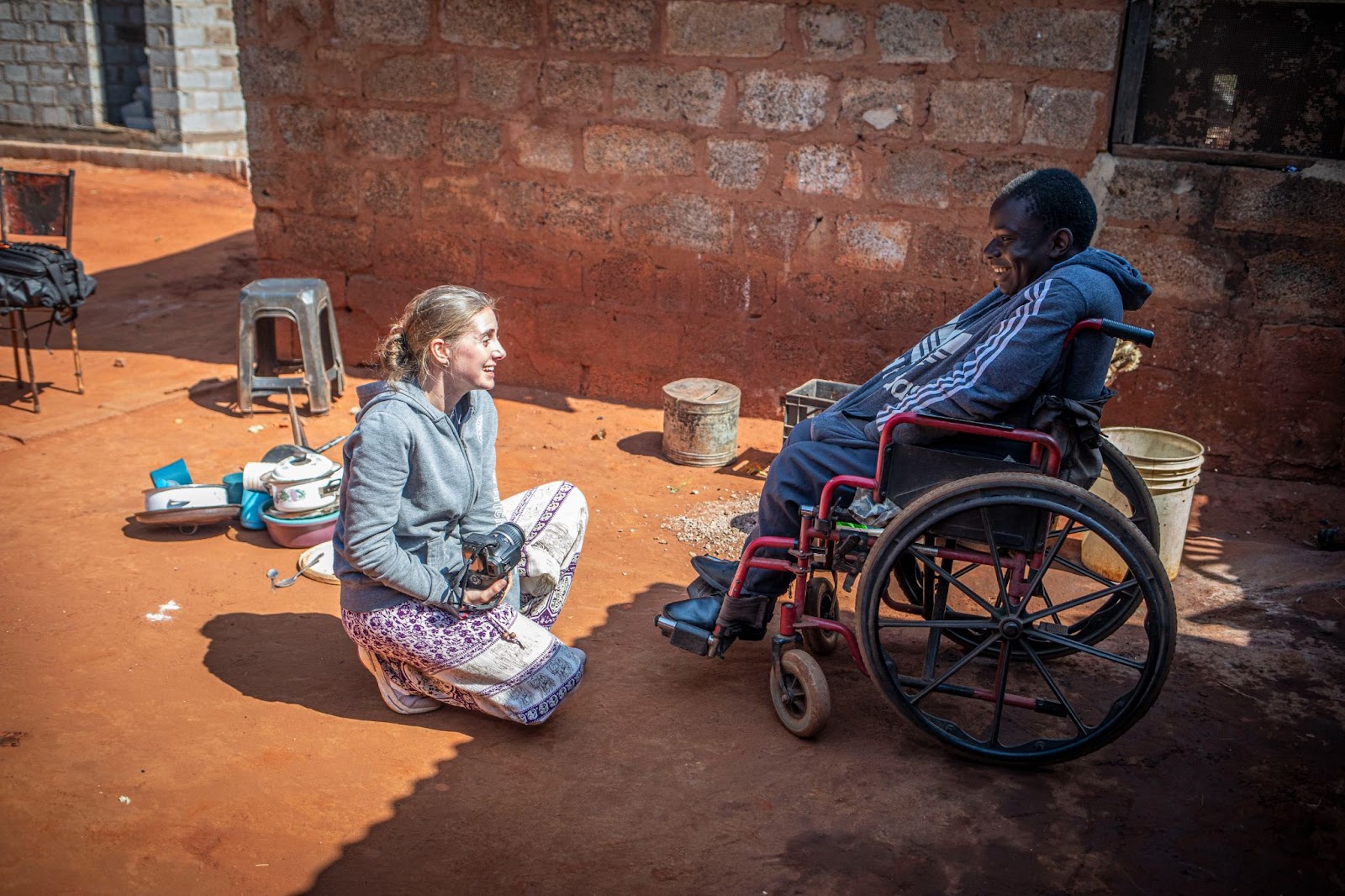 woman crouches on the ground, smiling and talking with a man in a wheelchair outside a brick building