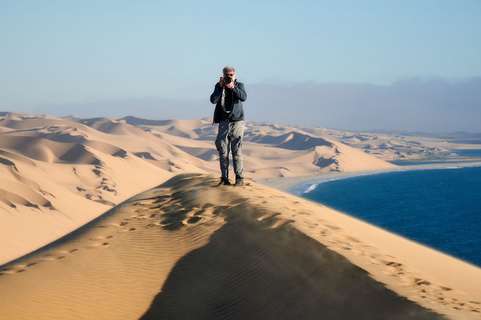 man stands on a sand dune, taking a photo, with rolling dunes