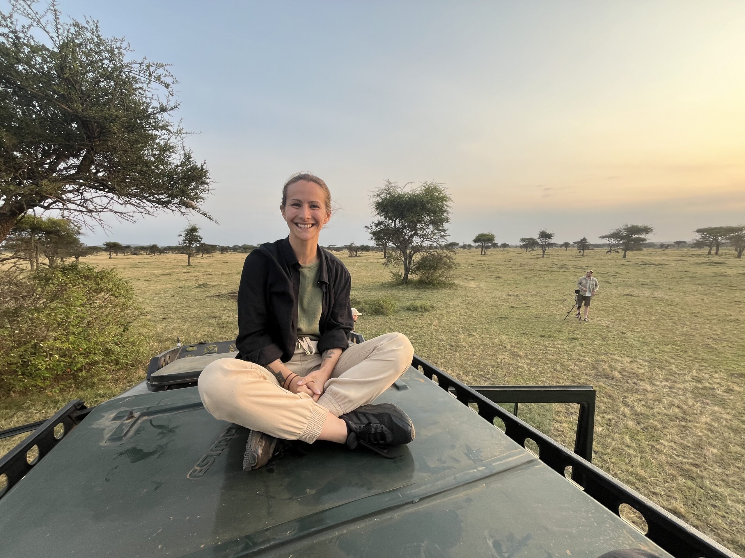 Smiling woman sitting cross-legged on top of a safari vehicle in an open savannah