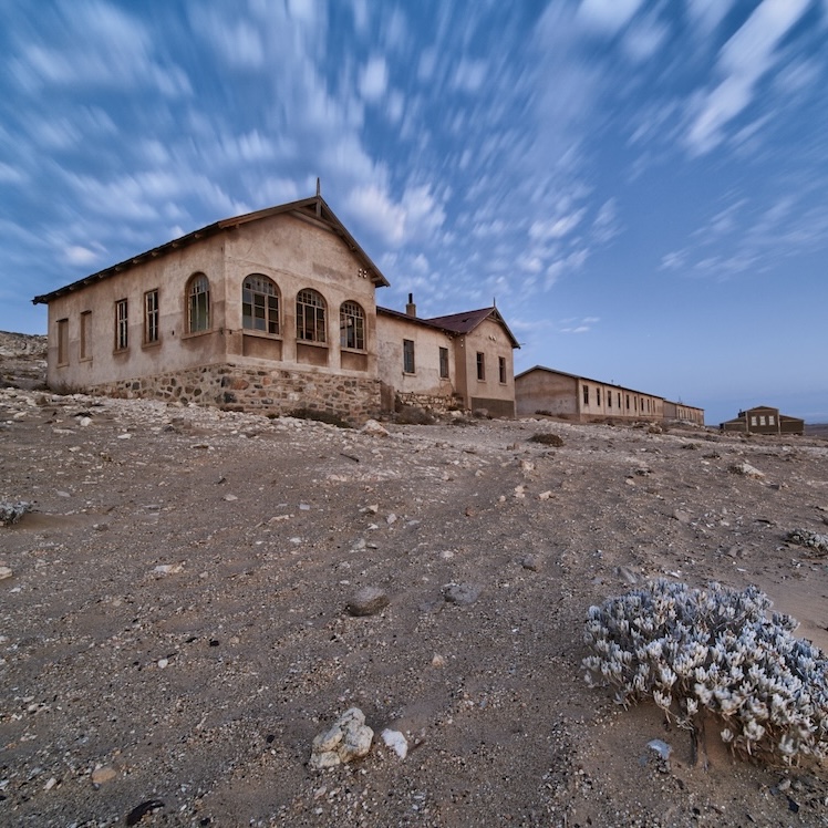 Abandoned buildings in a desert landscape under a cloudy blue sky