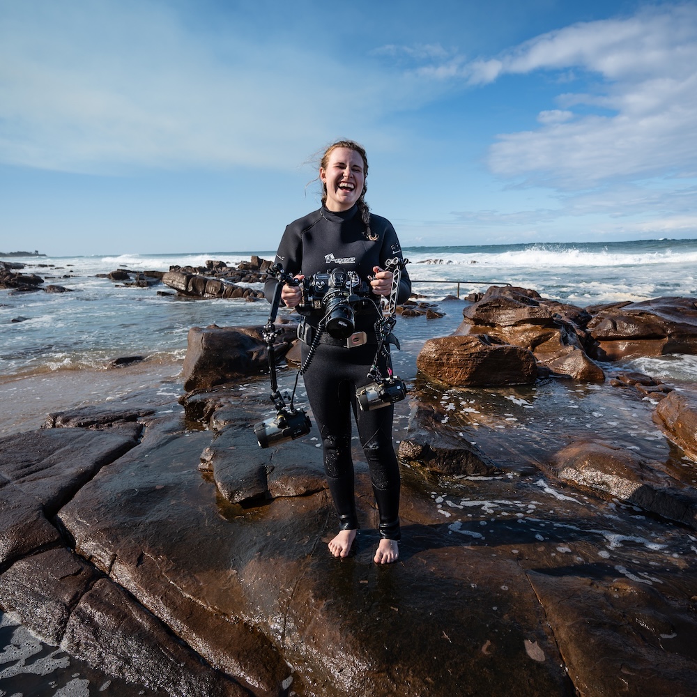 Woman in wetsuit holding underwater camera on rocky shore