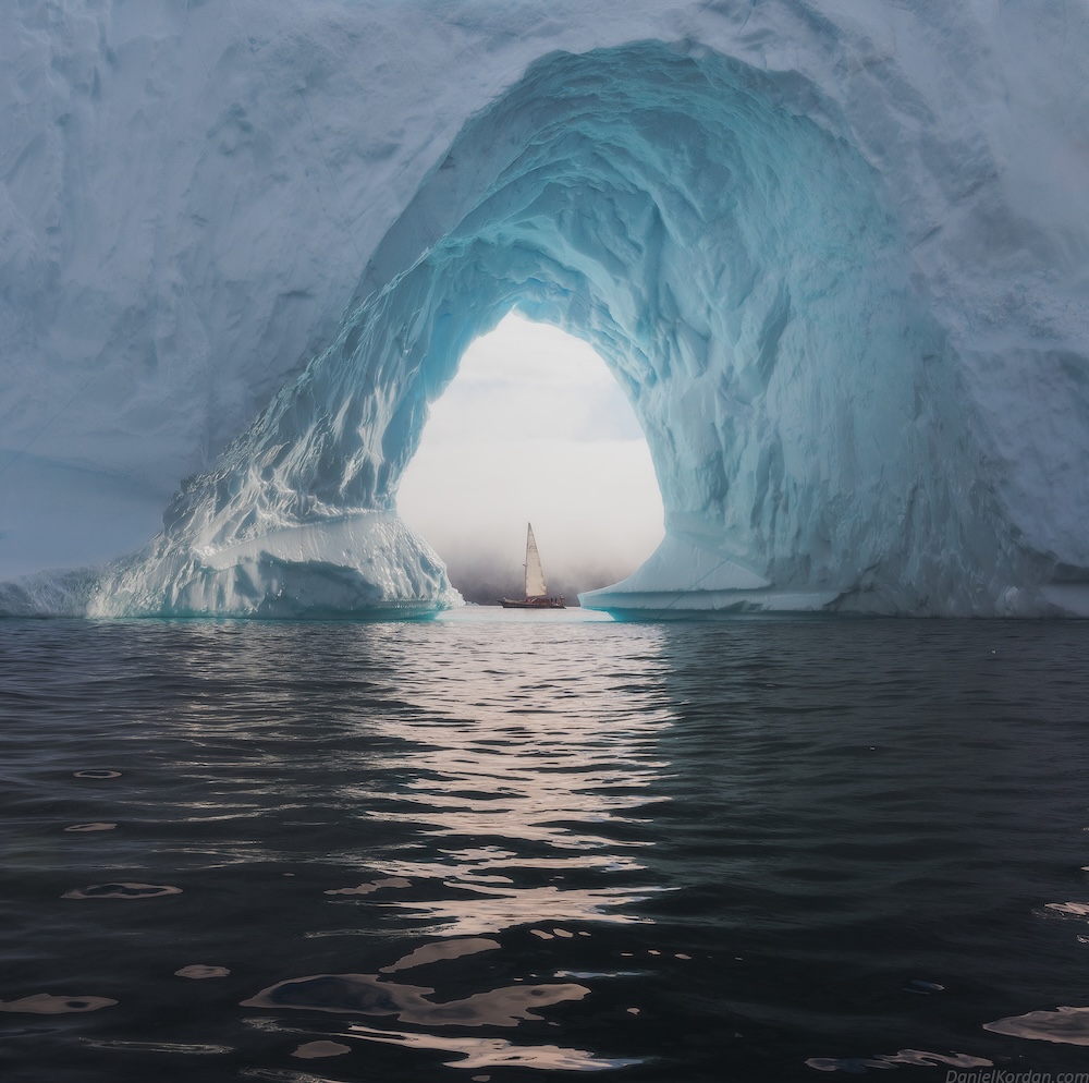 A photograph of a sailboat passing through a large ice arch in Greenland.