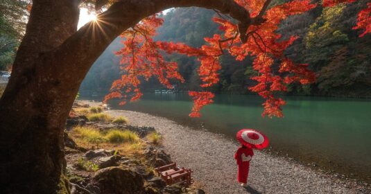 A person in a red kimono with a matching parasol stands beneath a radiant autumn tree by a tranquil river. Sunlight filters through fiery red and orange leaves, casting a warm glow over the serene Japanese landscape.