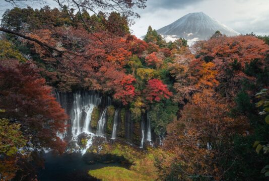 A waterfall cascades through vibrant autumn forest into a tranquil pool, with Mount Fuji rising snow-capped in the background. Red, orange, and yellow foliage surrounds the scene, blending dramatic natural beauty with iconic Japanese geography.