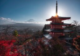 Red and white Japanese pagoda glows with sunlight behind its top tier, framed by fiery autumn foliage. Mount Fuji rises in the background, partially veiled by clouds, adding depth and elegance to the serene landscape.