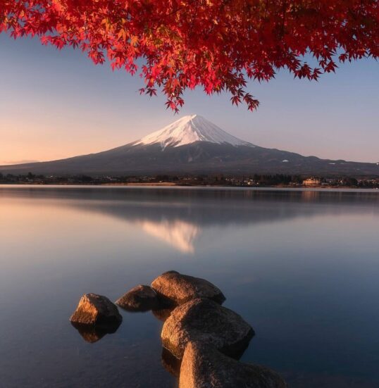 Mount Fuji’s snow-capped peak reflects in a calm lake, framed by vibrant red autumn leaves above. A natural stone path leads into the water, blending serene blues with fiery seasonal color in a harmonious Japanese landscape.