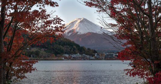 Mount Fuji glows under soft sunrise light, framed by brilliant red autumn leaves. A tranquil lake reflects the snow-capped peak and nearby village nestled at the mountain’s base, creating a serene, picturesque scene.
