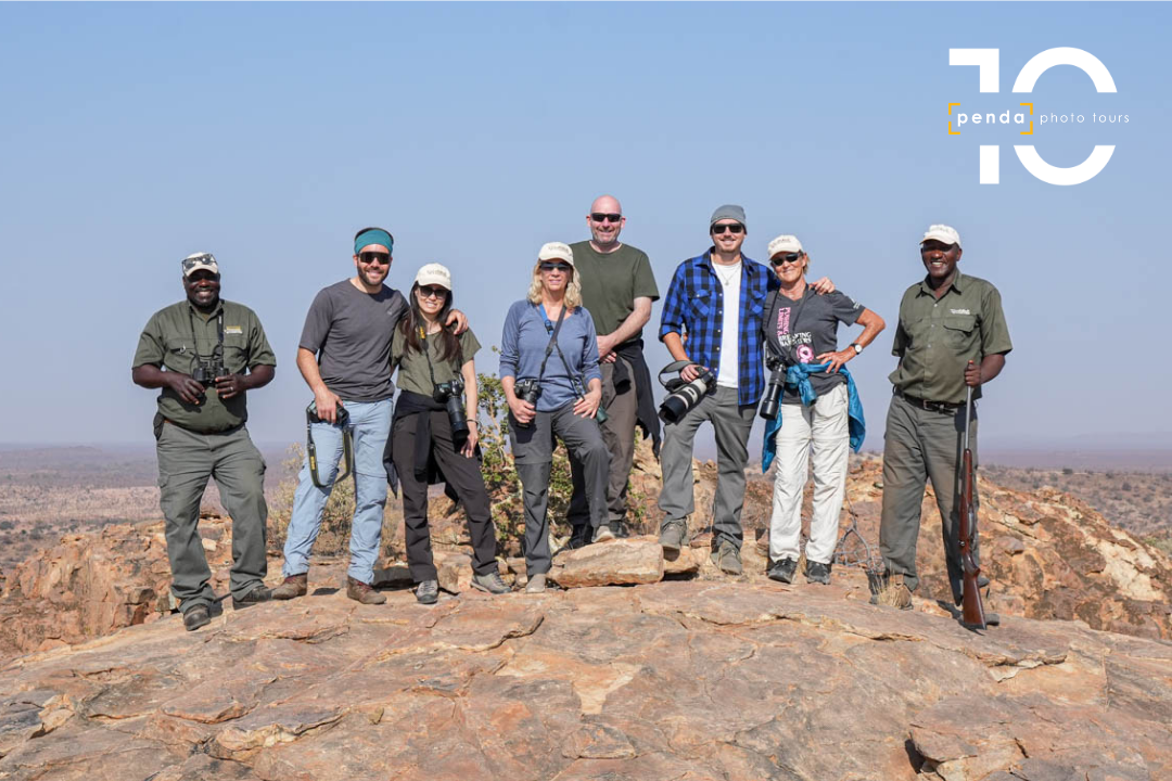 A group of guides and photographers pose together with cameras and binoculars atop a rocky outcrop overlooking the bush, marking Penda Photo Tours' 10th anniversary.