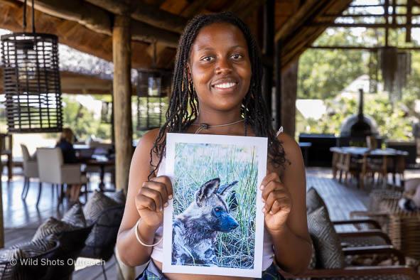 A smiling participant proudly holds a printed photograph of a wild dog beneath a thatched safari lodge, captured during the Wild Shots Outreach programme with Penda Photo Tours.