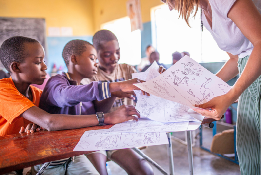 Three boys point at bird illustrations on coloring pages held up by a woman in a classroom, photographed on a Humanitarian Photography Workshop, Victoria Falls, by Anna Lusty.