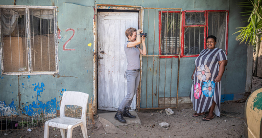 A photographer raises a camera to photograph a smiling woman standing in a floral striped dress outside a weathered, colorfully painted house, on a Humanitarian Photography Workshop, Victoria Falls, by Anna Lusty.