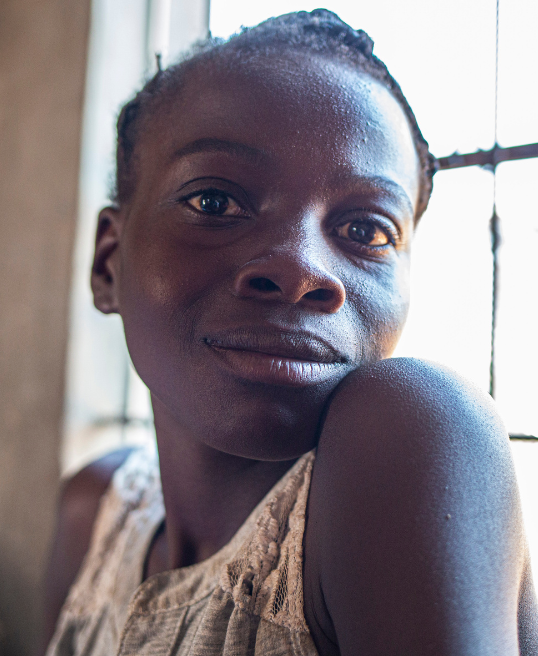 A girl rests her chin on her arm, gazing gently at the camera with soft window light on her face, photographed on a Humanitarian Photography Workshop, Victoria Falls, by Anna Lusty.