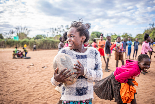 A girl smiles broadly while holding a worn leather ball, surrounded by other children playing on red dirt ground, photographed on a Humanitarian Photography Workshop, Victoria Falls, by Anna Lusty.