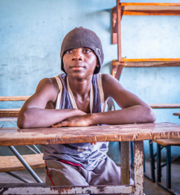 A boy in a beanie and tank top rests his arms on a wooden desk, looking calmly at the camera inside a classroom, photographed on a Humanitarian Photography Workshop, Victoria Falls, by Anna Lusty.